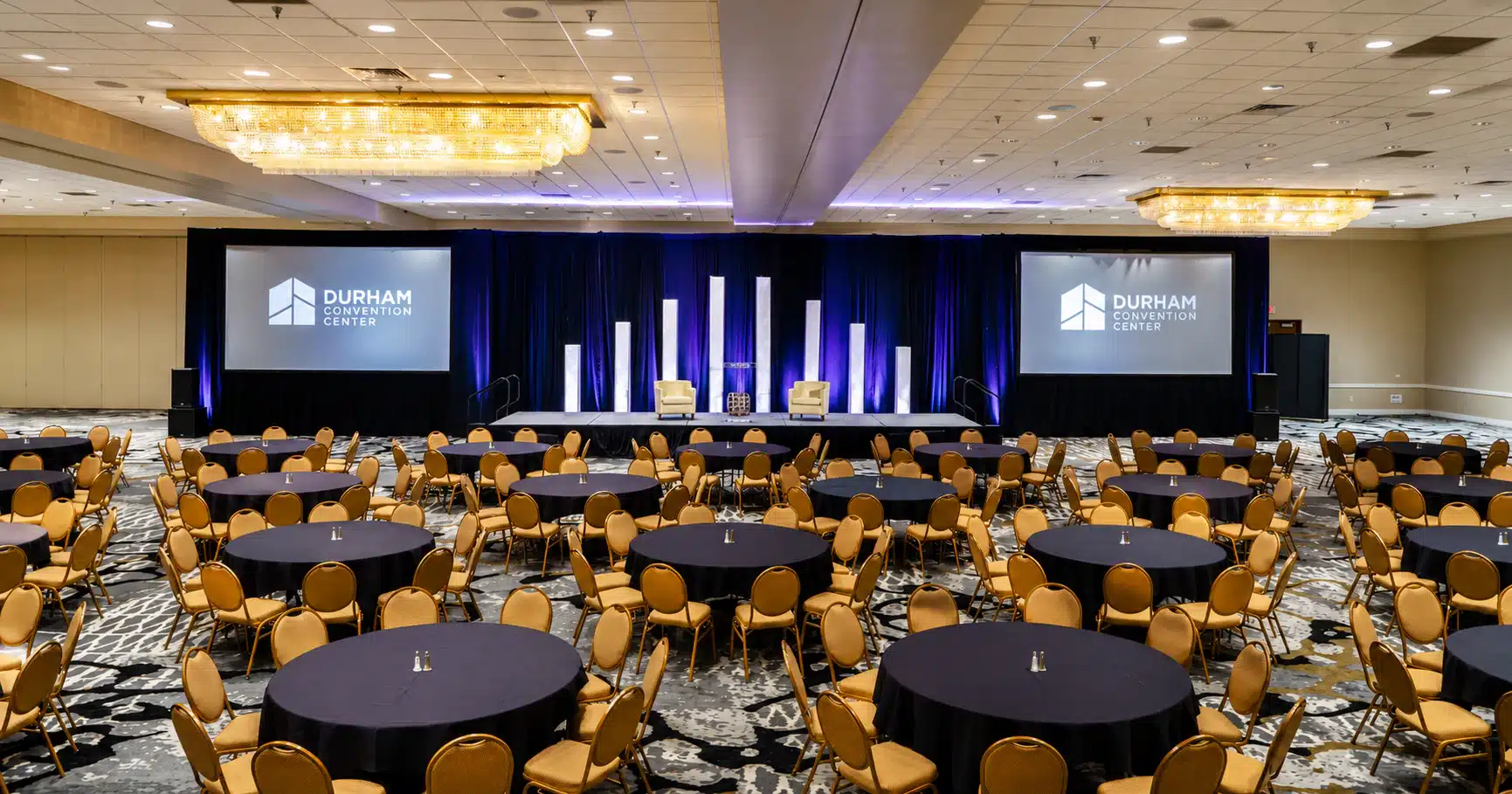 durhamconvention Conference hall with round tables and gold chairs facing a stage with blue curtains and two screens showing Durham Convention Center logo.
