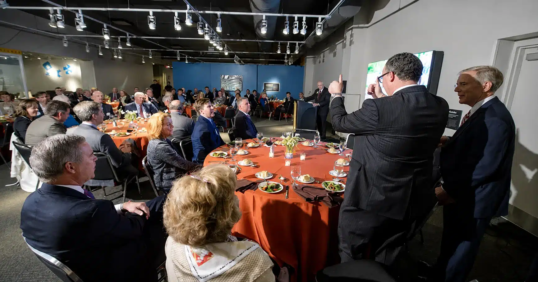 lifeandscience Large group of people in business attire seated around round tables with orange tablecloths, listening to a speaker at a podium.
