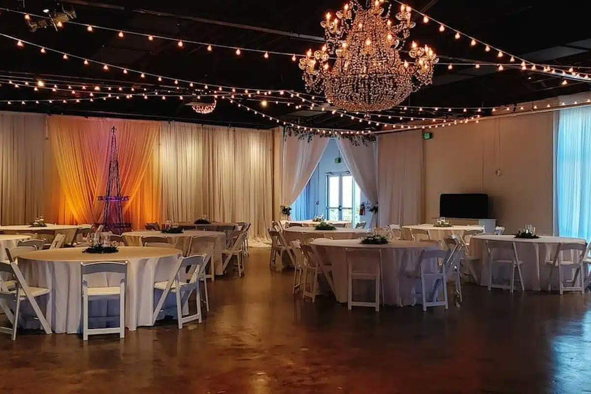 the-fairview-1200×800 Elegant event hall with round tables in white linen, white chairs, draped curtains, and a crystal chandelier.