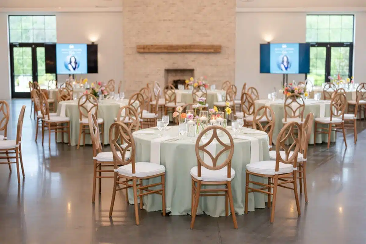 the-maxwell-1200×800 Banquet hall set for an event with round tables, pale green tablecloths, floral centerpieces, and wooden chairs with white cushions.