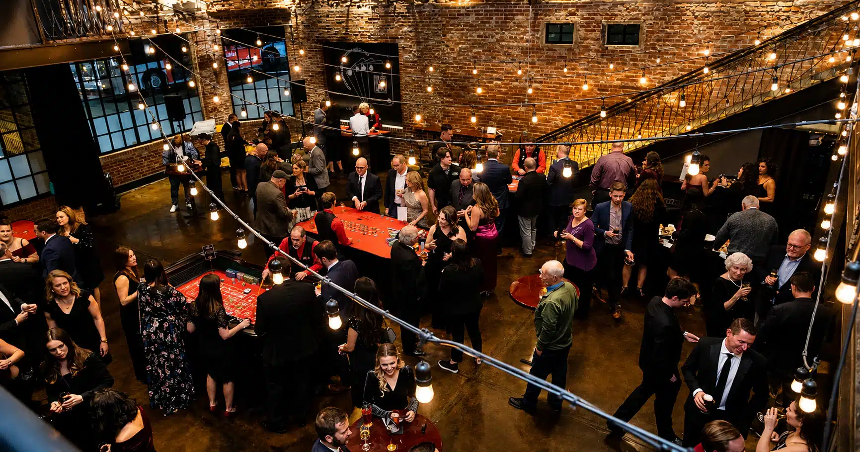 thecookery Crowd of well-dressed guests at a brick-walled venue with string lights, mingling around red casino tables at a social event.
